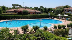 A swimming pool with a circular design, surrounded by lounge chairs and umbrellas, with buildings and greenery in the background.