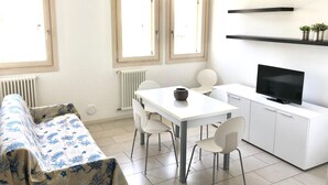 A modern, well-lit room with a sofa, dining table, chairs, a TV, and a kitchen area with a shelf.