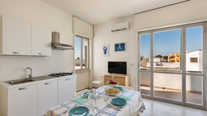 Cocina moderna con estufa a gas, lavamanos y armarios blancos. Se aprecia una mesa de comedor puesta con platos y vasos, y un televisor al fondo.