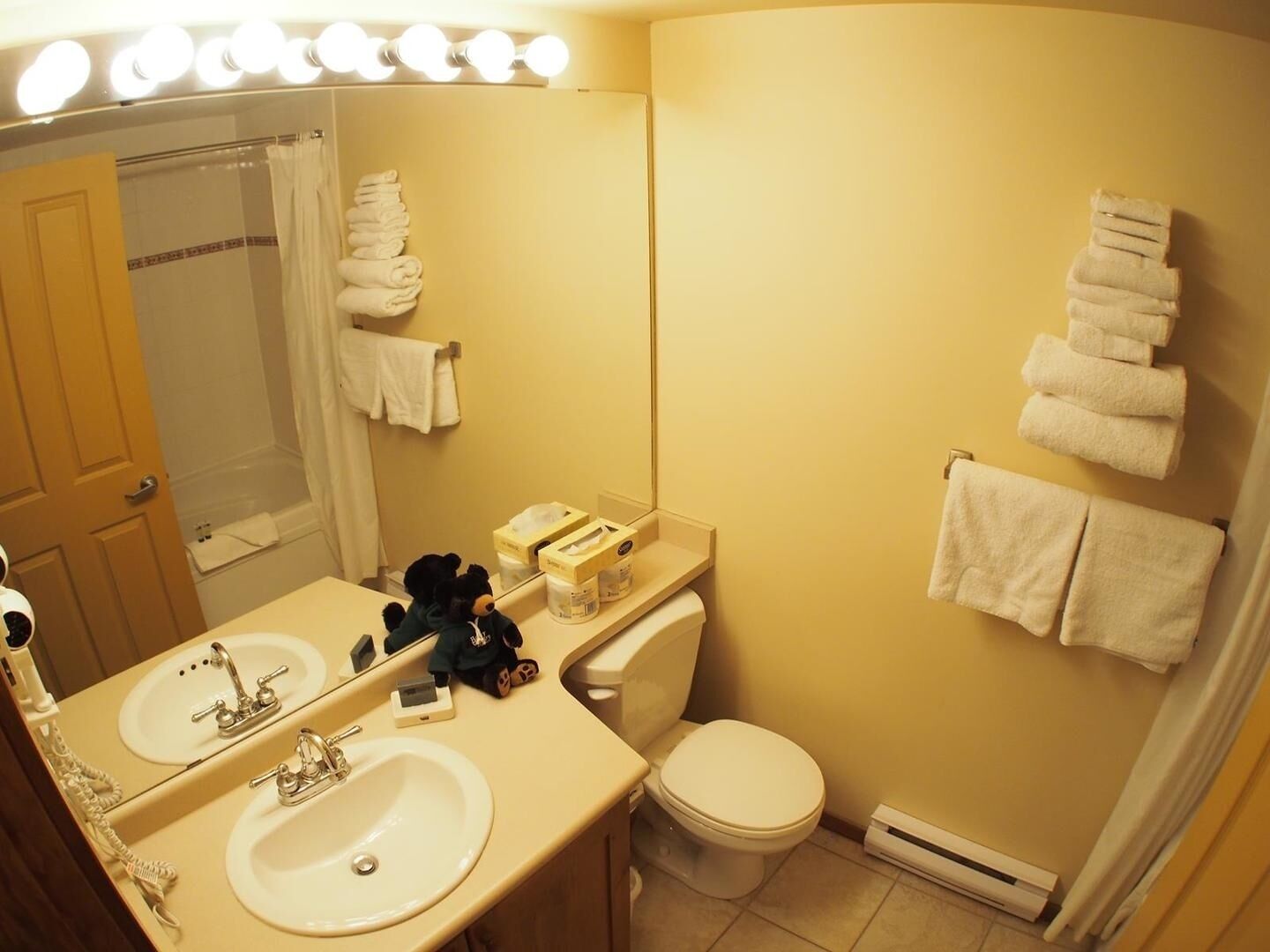 A bathroom with two sinks, a toilet, a mirror with lights, and towels on the wall.