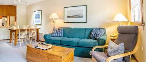 A cozy living room with a teal sofa, wooden coffee table, and a grey armchair. There is a dining area with a wooden table and chairs, and a kitchen in the background.