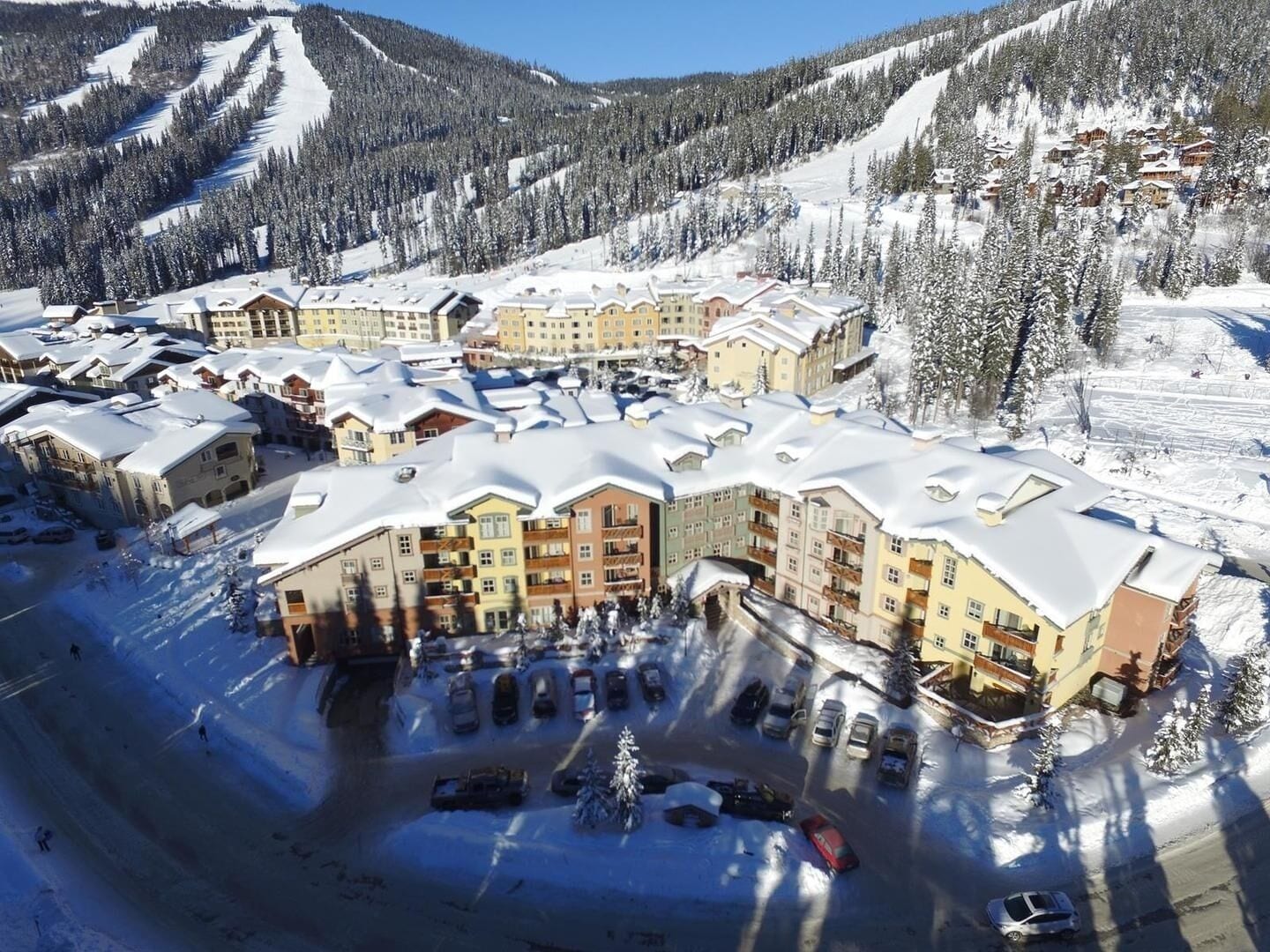 A snow-covered resort with multiple buildings, a parking lot, and a ski slope in the background.