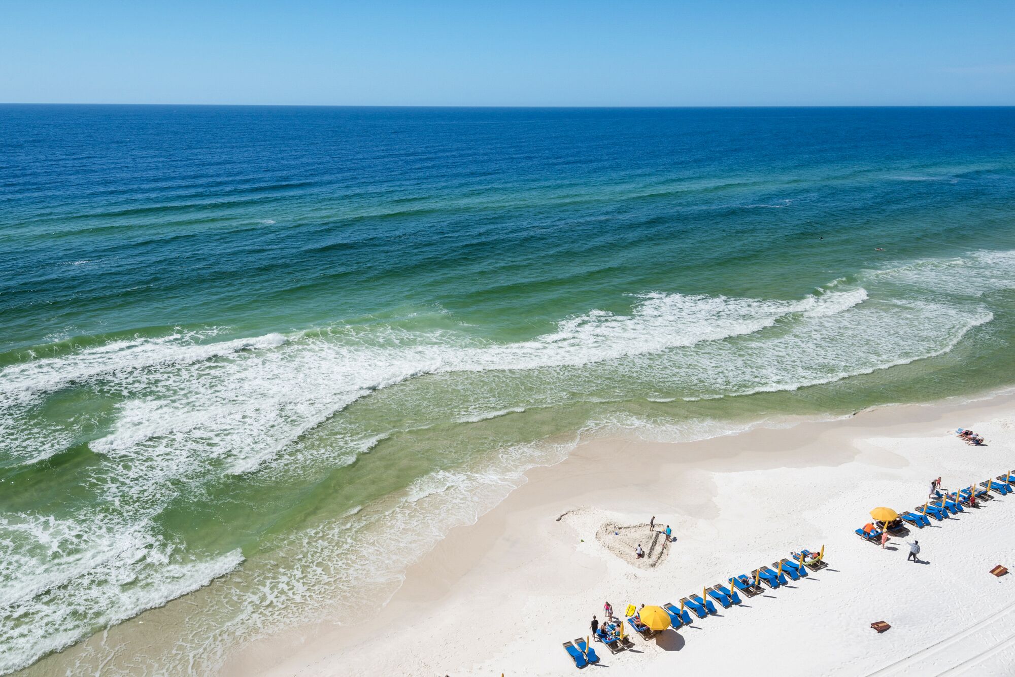 A beach with clear blue water, a sandy shore, and rows of blue beach chairs and yellow umbrellas.