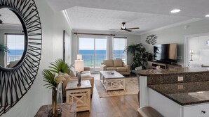 A spacious living area with a large window overlooking the ocean, a ceiling fan, and a modern kitchen with granite countertops. - Treasure Island 1003 (Panama City Beach)