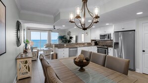 A spacious kitchen with white cabinets, a stainless steel refrigerator, and a dining area with a wooden table and chairs. - Treasure Island 1003 (Panama City Beach)