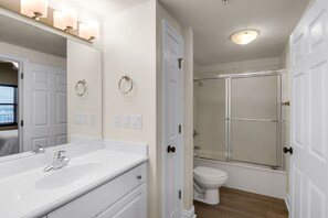 A bathroom with a white sink, mirror, and toilet. There is a shower enclosure and a door with a towel ring. - Treasure Island 1003 (Panama City Beach)