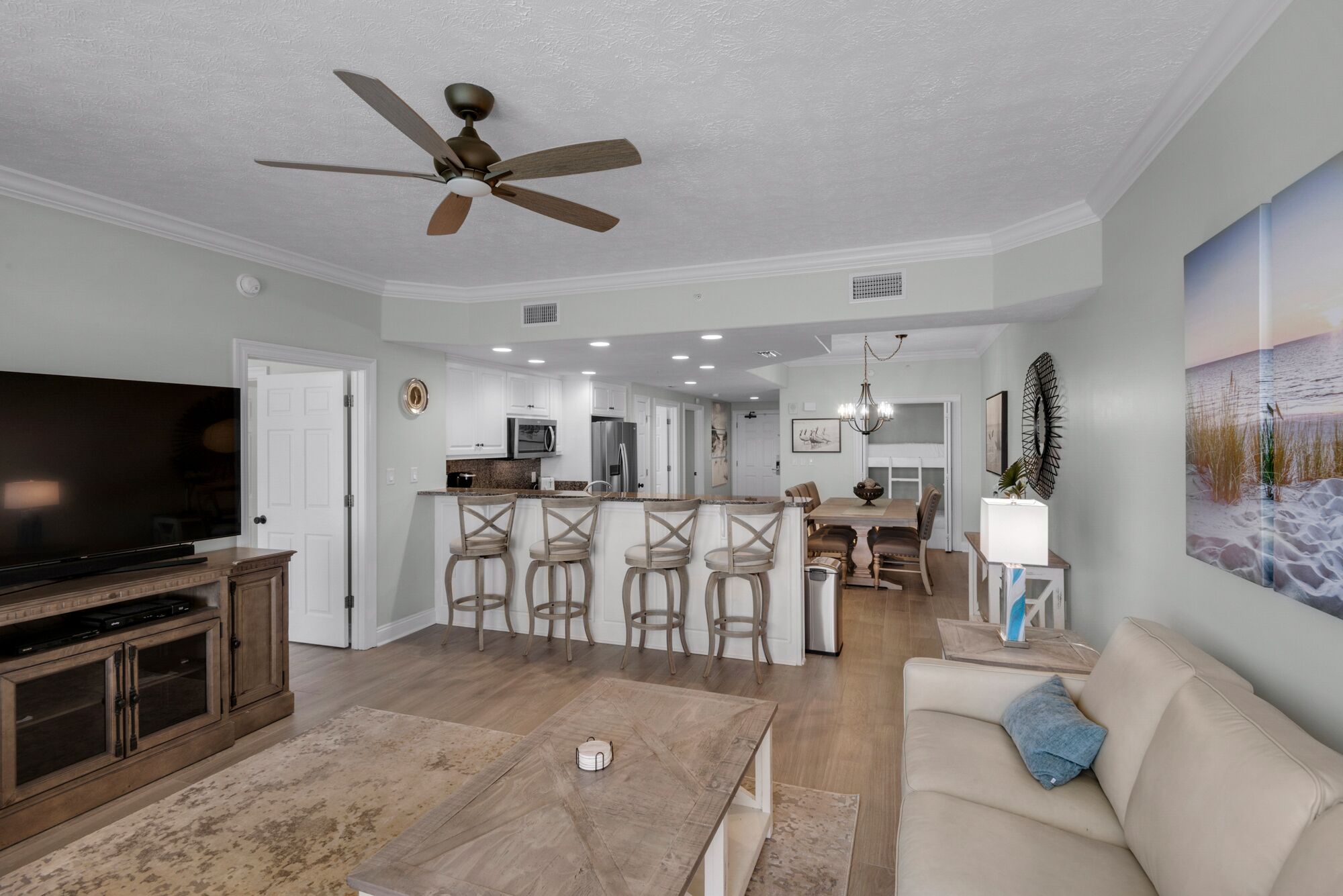 A modern living room with a TV, a ceiling fan, a dining area with bar stools, and a kitchen in the background.