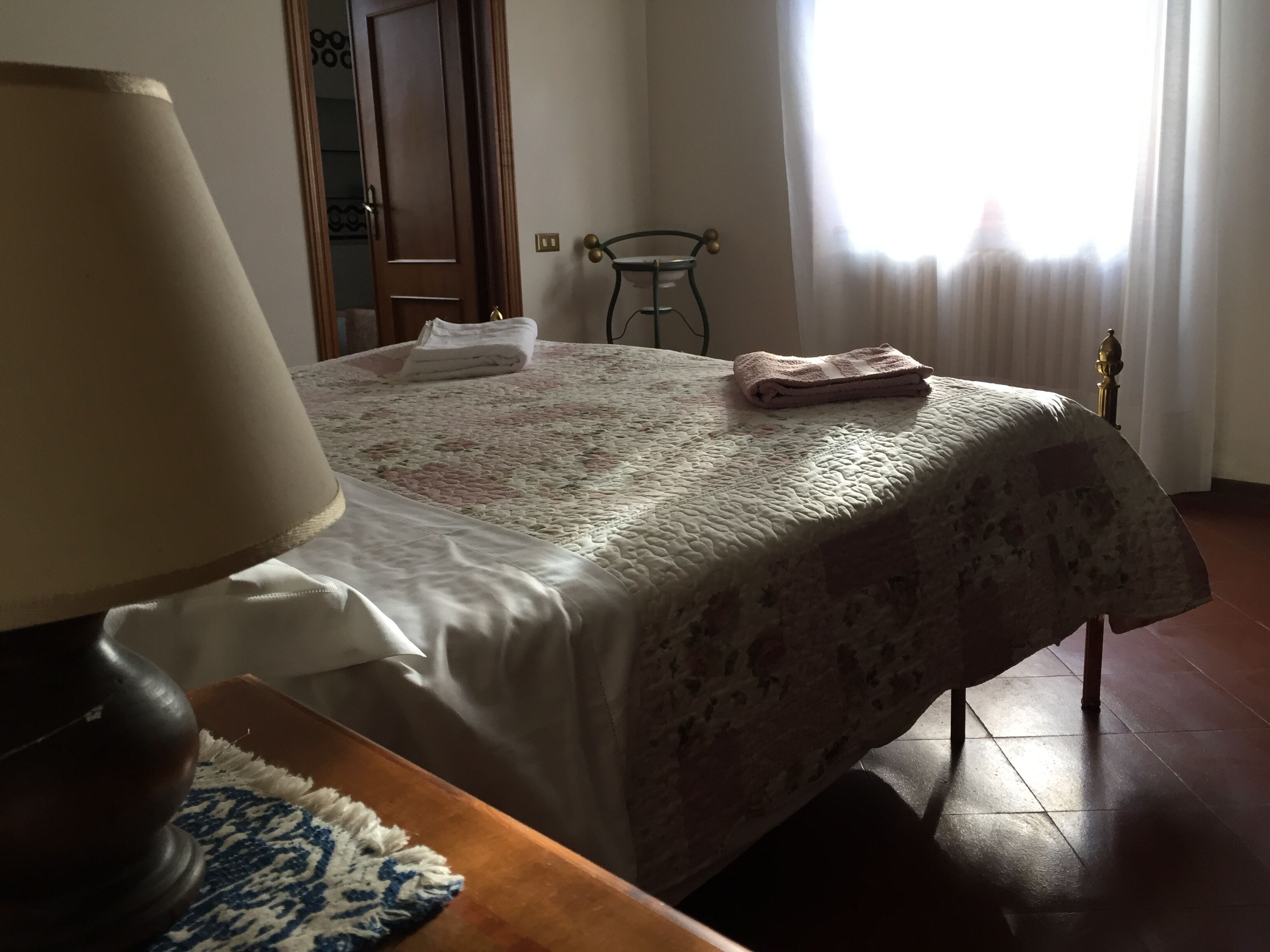 A neatly made bed with a floral bedspread and two towels, a wooden nightstand with a lamp, and a window with curtains.