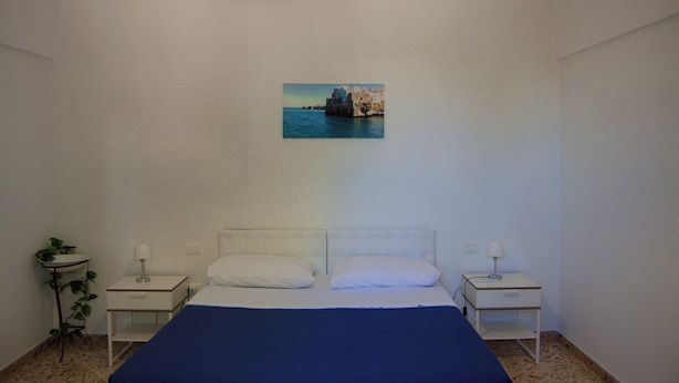 A bedroom with a bed, two bedside tables, a nightstand, a wall-mounted picture, and a small potted plant.
