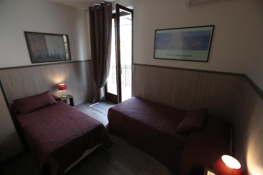 Ein Hotelzimmer mit zwei Betten, einem Nachttisch mit Lampe, einem gerahmten Bild an der Wand und einem Fenster mit Vorhängen.