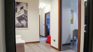 A hallway with a wooden floor, a white bench, and a red chair. There is a framed picture on the wall and a door on the right.