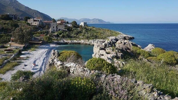 Uma vila costeira com edifícios de pedra, uma praia rochosa e vista clara para o mar.