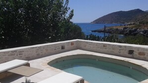 Uma piscina infinita de pedra com uma espreguiçadeira branca e uma mesa pequena, rodeada por um muro de pedra com vista para o mar e uma colina ao fundo.