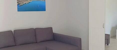 A modern living room with a grey sectional sofa, a white wardrobe, and a painting of a coastal landscape.