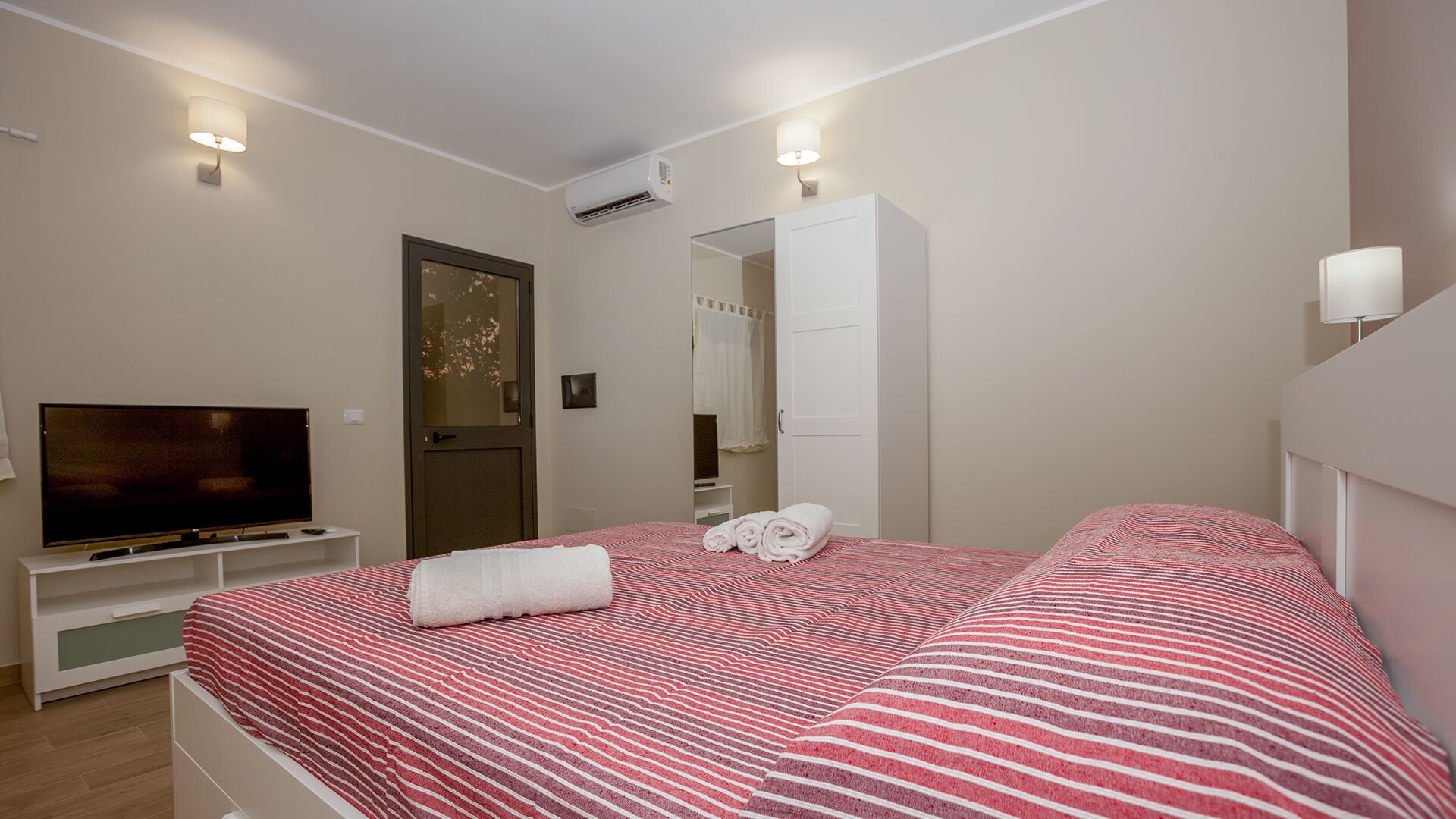 Een slaapkamer met een televisie, een bed met gestreepte beddengoed, een witte garderobe en een badkamers deur.