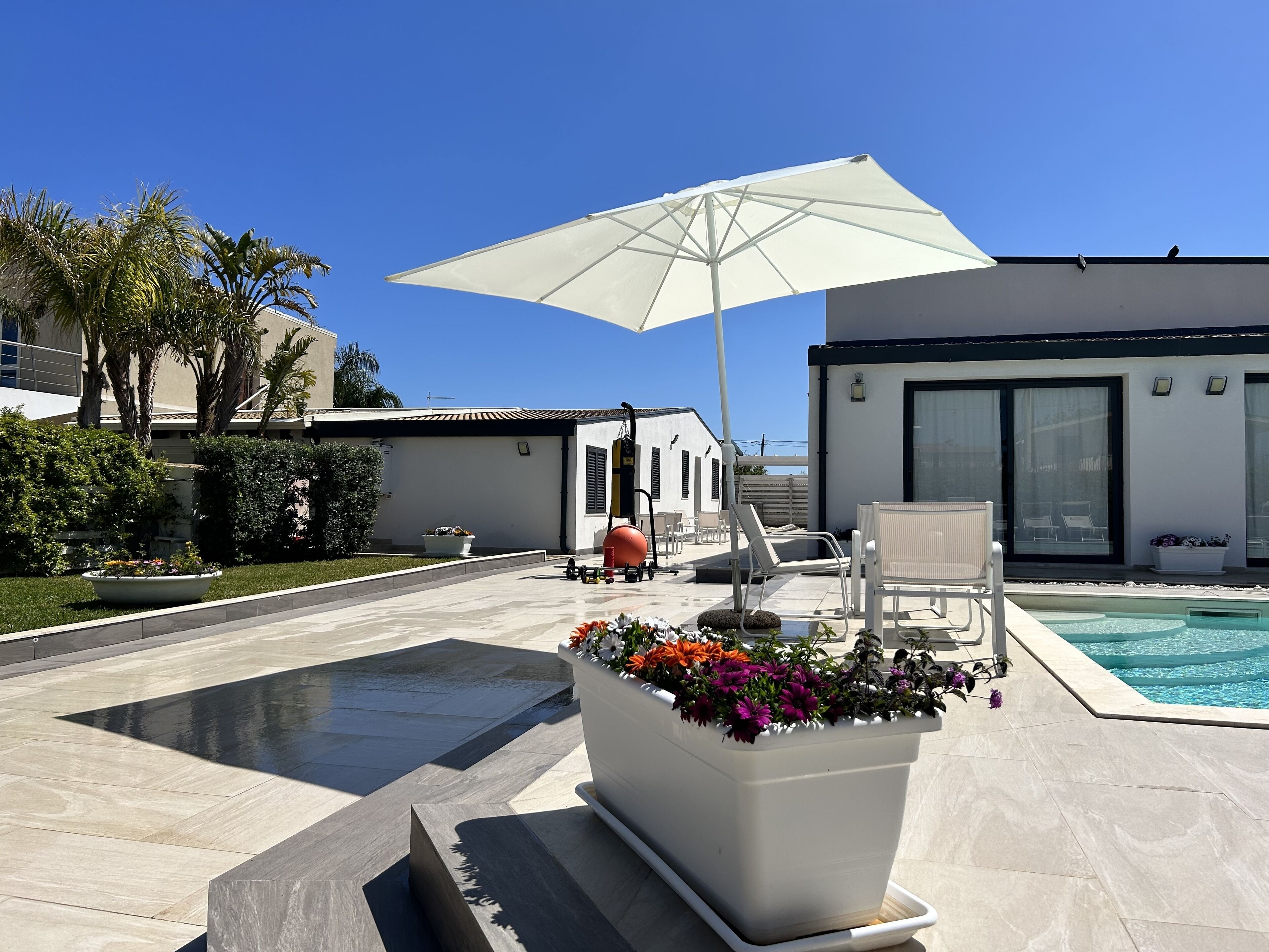 A modern outdoor pool area with a white umbrella, lounge chairs, and a flower planter.
