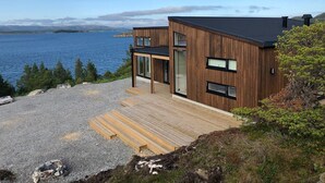 A modern two-story house with a wooden exterior, large windows, and a deck leading to a gravel area. - New holiday home, idyllically located by the sea (Kolvereid)