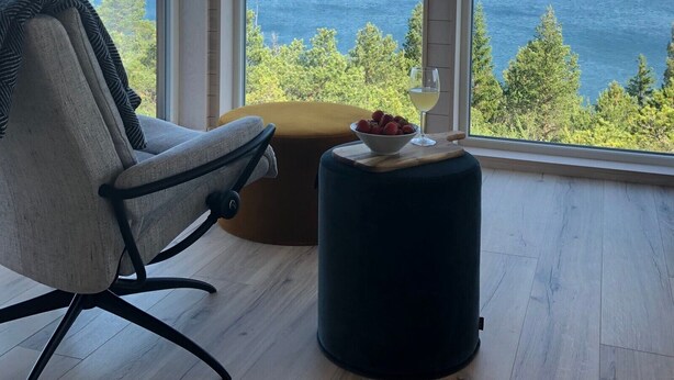 A room with a large window offering a view of a body of water and greenery. There is a chair and a small round ottoman with a bowl of fruit and a glass of wine on the windowsill. - New holiday home, idyllically located by the sea (Kolvereid)