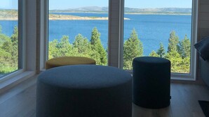 A room with large windows offering a view of a body of water and distant land. There are two round ottomans on the wooden floor. - New holiday home, idyllically located by the sea (Kolvereid)