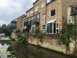 A multi-story brick building with balconies, large windows, and a waterway in front. - River Courtyard Apartment - St Neots (Saint Neots)