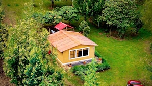 Une maison jaune à un seul étage, avec un toit rouge, entourée d’une végétation luxuriante et une voiture rouge garée à proximité.