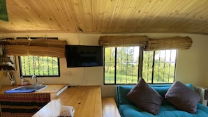 Une pièce avec un plafond en bois, un téléviseur fixé au mur, une fenêtre donnant sur un paysage verdoyant, un canapé turquoise agrémenté de coussins et une table en bois sur laquelle se trouvent une calculatrice et un tissu bleu.
