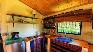 Une cuisine compacte équipée d’un réfrigérateur, d’une cuisinière et d’un évier. On y trouve des étagères au-dessus du plan de travail et une fenêtre avec des stores.