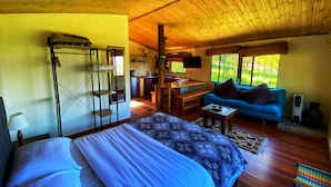 Une pièce chaleureuse avec un lit, un canapé bleu, un plafond en bois et un coin cuisine visible à travers une porte ouverte.