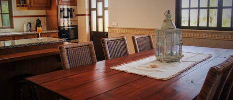 A wooden dining table with a glass lantern centerpiece, surrounded by wicker chairs, in a kitchen with wooden cabinets and a window with a view.