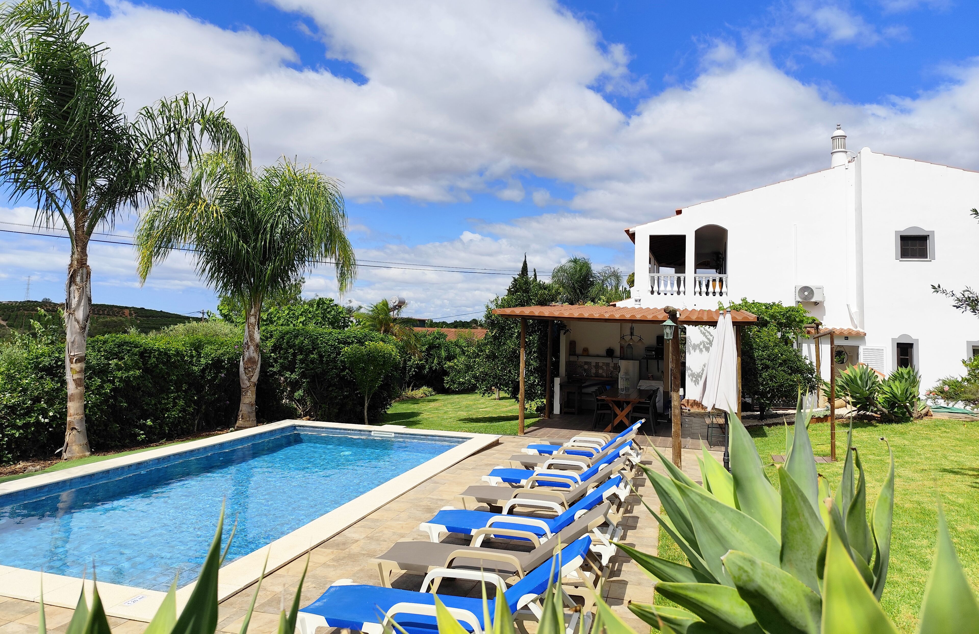 A swimming pool with lounge chairs, a white building with a balcony, and surrounding greenery.