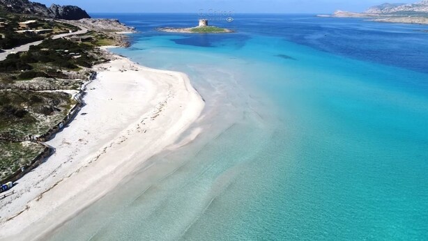 Ett flygbildsbild av ett kustområde med en sandstrand, klart blått vatten och en liten ö med ett torn.