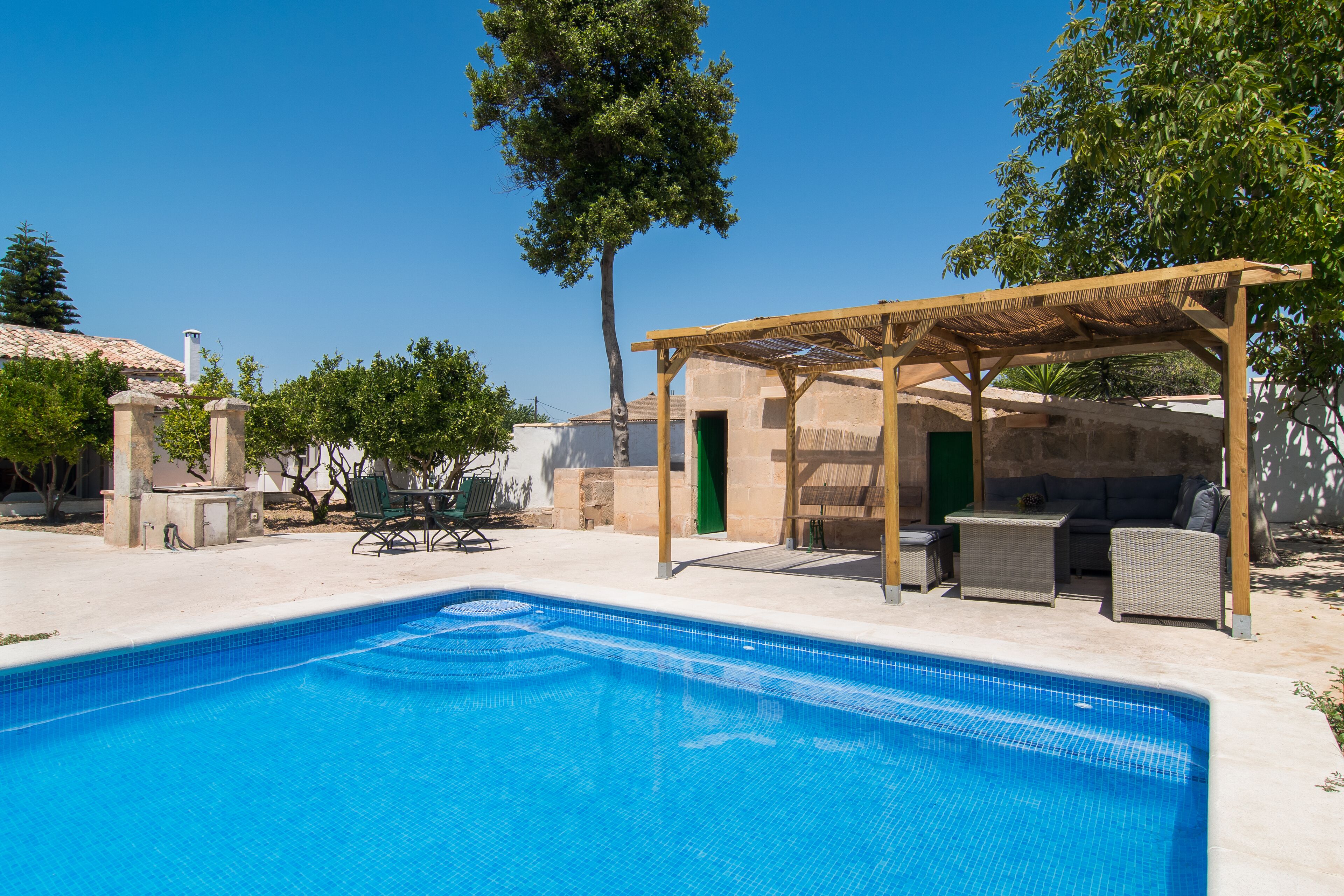 Una piscina con un pérgola y mobiliario exterior, rodeada de árboles y un edificio con techo de tejas.