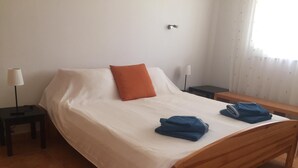 Een net opgemaakt bed met een witte dekbedovertrek en een oranje kussen, twee opgevouwen handdoeken op het bed, een houten nachtkastje met een lamp, en een houten bankje naast het bed.