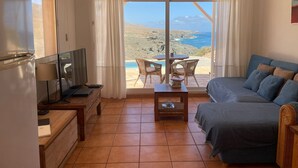 Een kamer met een bed, een televisie, een tafel met stoelen en uitzicht op zee.