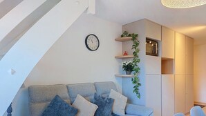 Ein Wohnzimmer mit einer Couch, einer Uhr, Pflanzenregalen und einem Essbereich mit Stühlen.