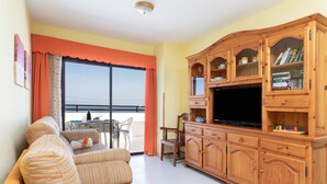 Een woonkamer met een bank, televisie, een houten kast en een balkon met een tafel en stoelen.