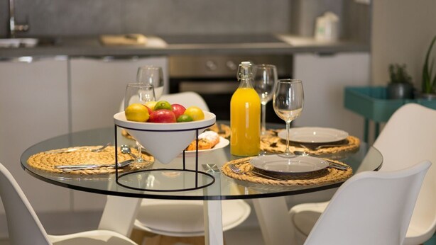 Et moderne spiseområde med et glasbord, hvide stole, en skål med frugt, en appelsinjuiceflaske og et vinglas.