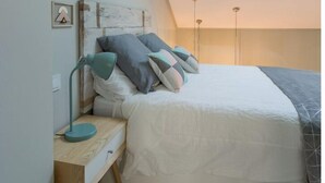 Et soveværelse med skråvæg, ovenlys, seng med puder, natbord med lampe og et lille billedramme på væggen.