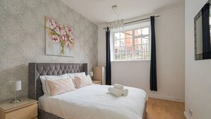 Ein Schlafzimmer mit einem großen Bett, einem Nachttisch, einer Lampe, einem Blumenbild, einem Fenster mit Vorhängen und einem Spiegel.