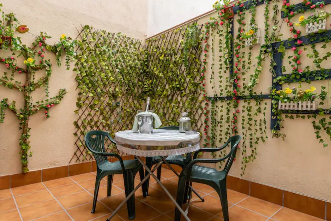 A small outdoor patio with a round table and chairs, surrounded by a lattice wall covered with climbing plants and flowers.