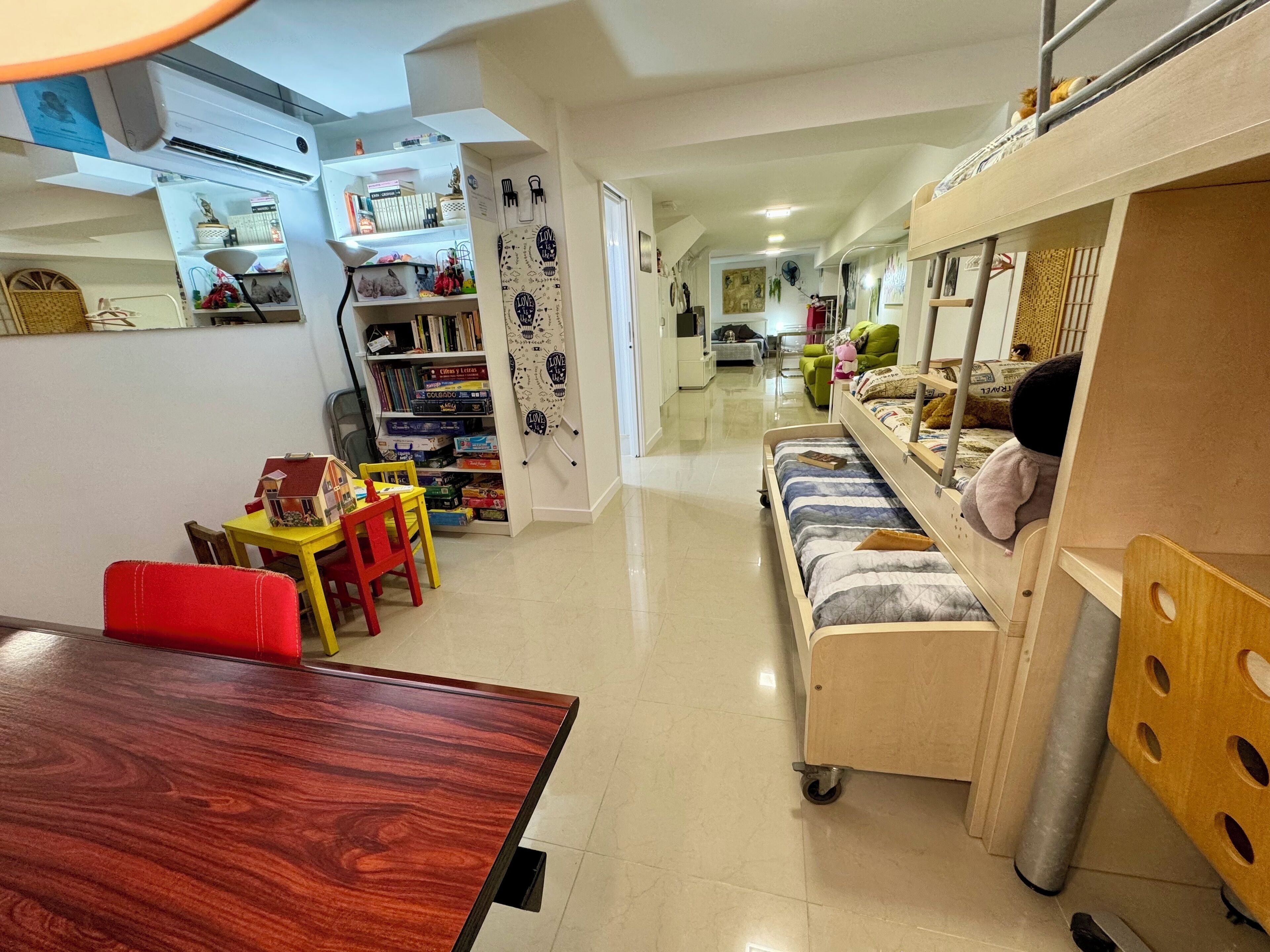Une pièce avec une table à manger en bois, un canapé avec des coussins, une bibliothèque et un lit superposé avec des jouets.