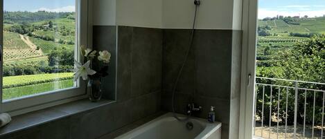 A bathroom with a bathtub, a shower, and a window with a view of a vineyard.