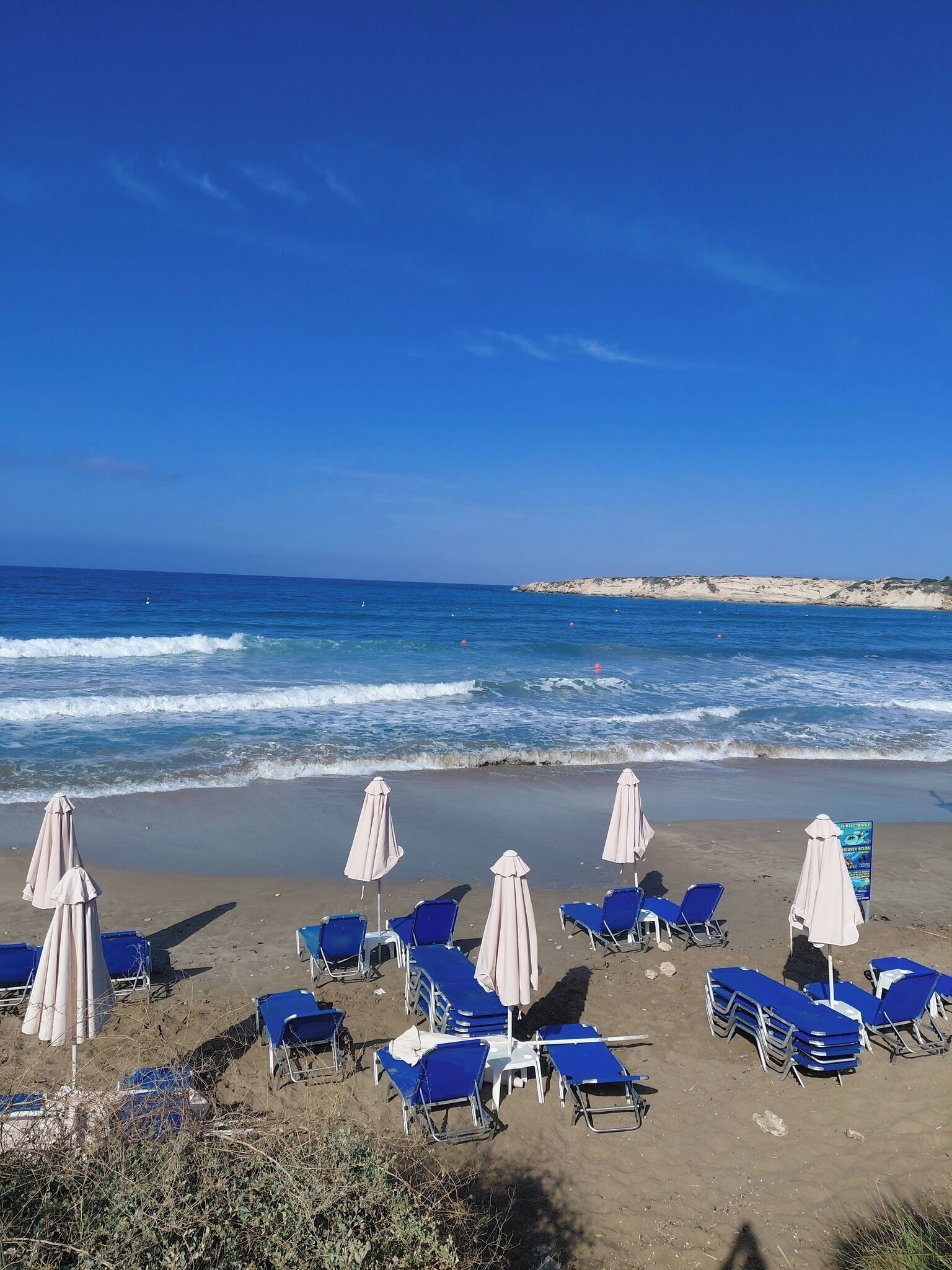 Una playa con hileras de sillas azules y sombrillas blancas, una orilla arenosa y el océano al fondo.