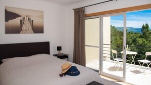 A bedroom with a bed, a nightstand, a lamp, a hat, and a view of a balcony with chairs and a table. - Spacious & new holiday home for max. 6 people (Aljubarrota)
