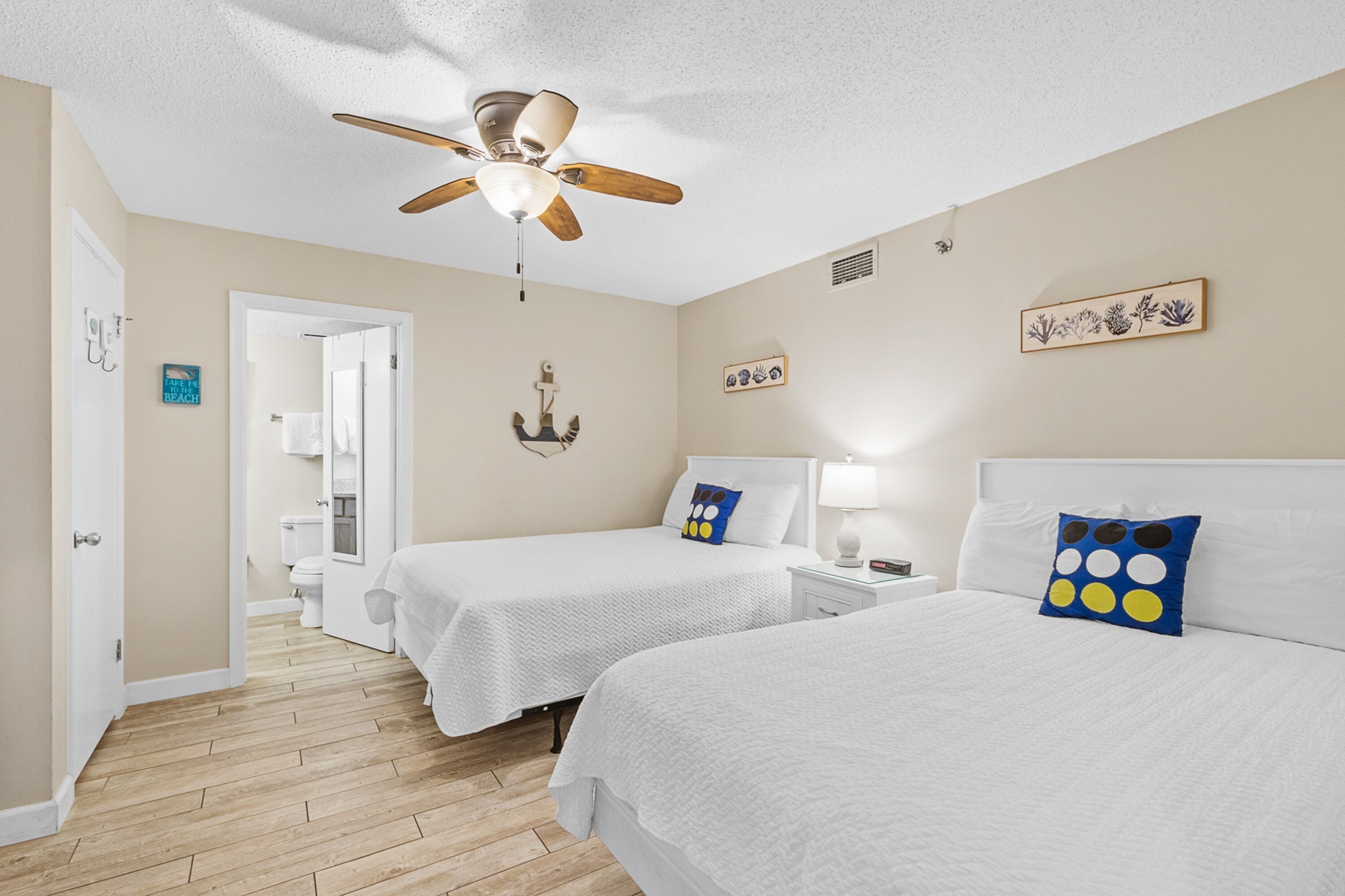 A hotel room with two beds, a ceiling fan, a bathroom with a toilet and sink, and a door leading to another room.