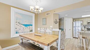 Un comedor con una mesa y bancos de madera, un cuadro de una escena de playa y una cocina con refrigerador y microondas.