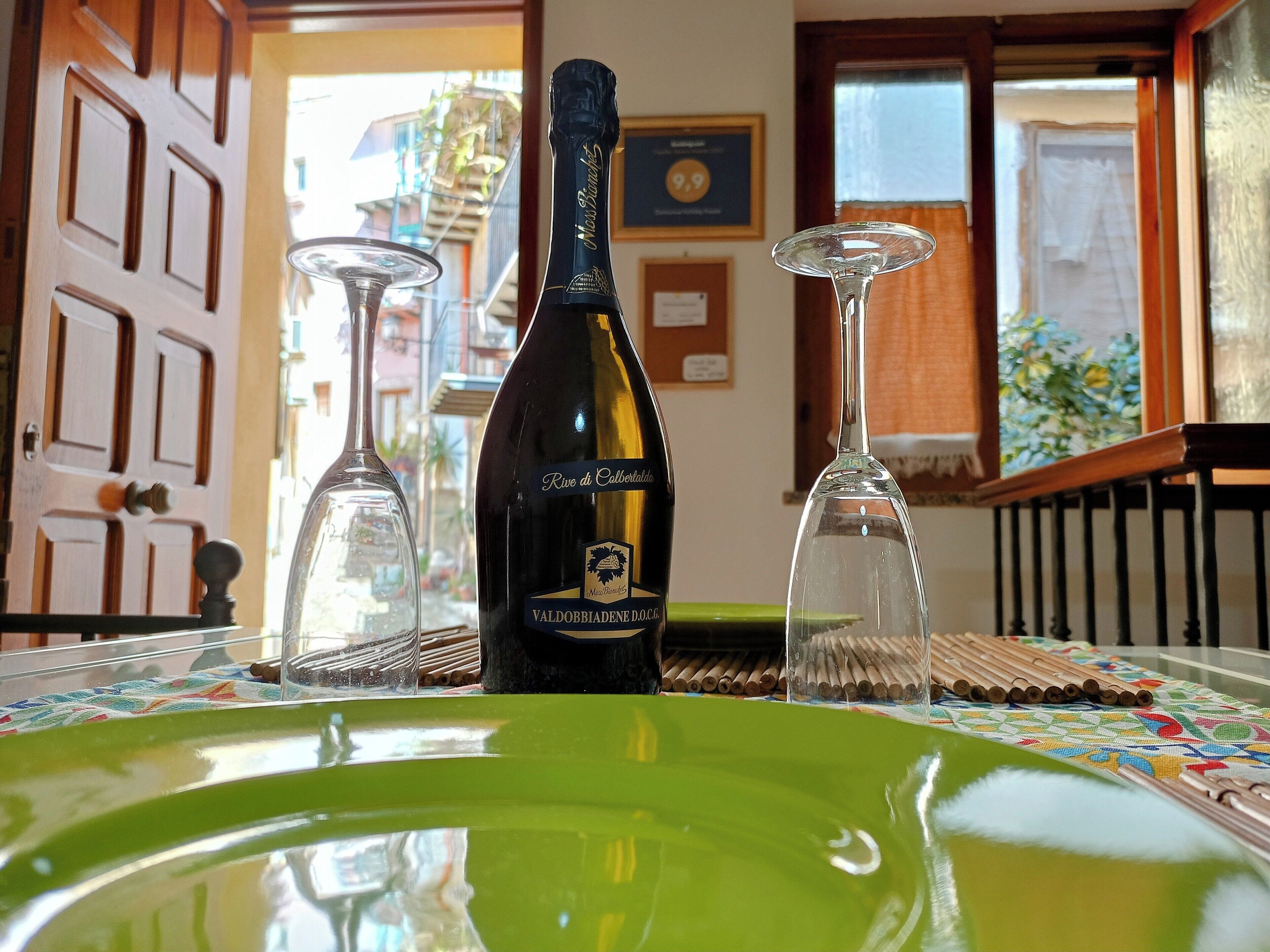 Eine Weinflasche, zwei Weingläser und ein grüner Teller auf einem Tisch mit gemustertem Tischläufer.