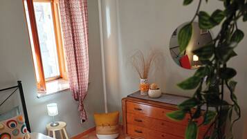 Ein Schlafzimmer mit Bett, Kommode, Fenster mit Vorhängen, Nachttischlampe und einem Blumentopf.