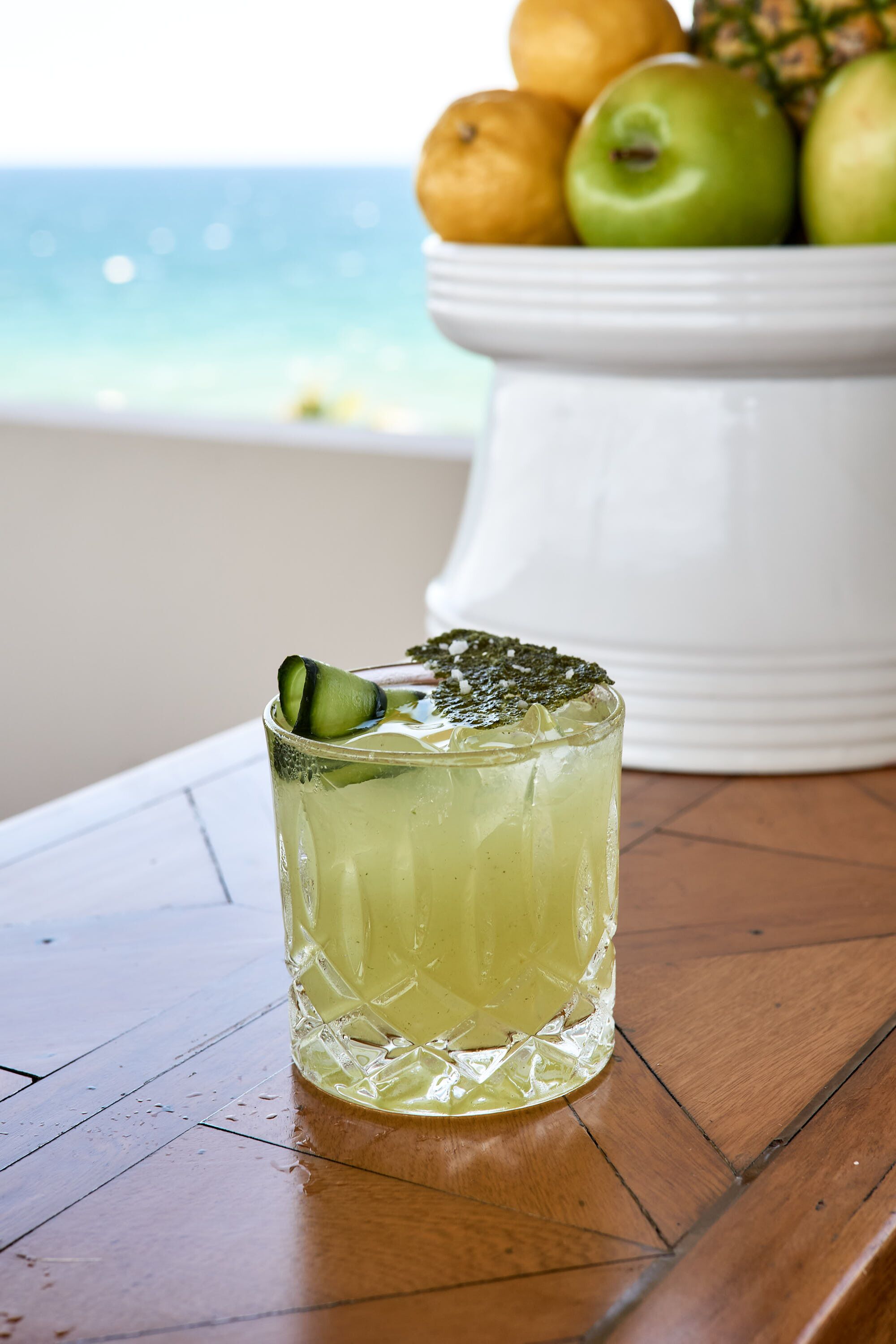 Un verre d’une boisson claire, avec une tranche de citron vert, posé sur une table en bois. En arrière-plan, on aperçoit un saladier rempli de fruits et une vue sur l’océan.