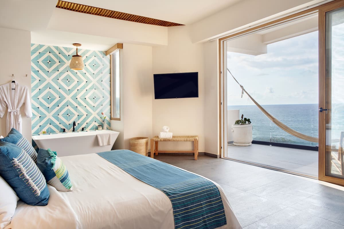 A modern bedroom with a bed, a TV, a bathroom, and a balcony with a view of the sea.